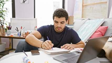 A focused man studying indoors with a notebook, laptop, and smartphone on a desk surrounded by art...
