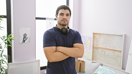 A confident young man with arms crossed and headphones stands in a bright art studio interior.