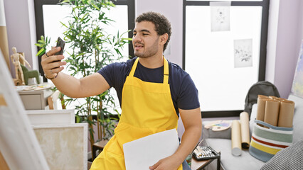 A cheerful man in a yellow apron takes a selfie in an art studio filled with plants and art...