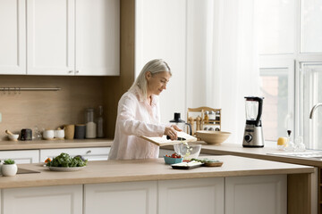 Pretty blonde senior woman chopping fresh vegetables on board on kitchen table, throwing slices...
