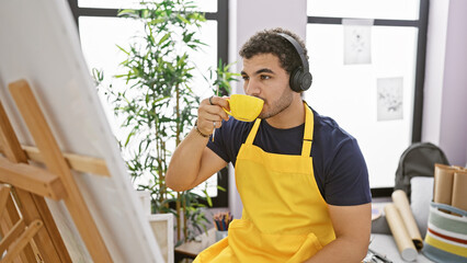 A young man with headphones sipping coffee in a bright art studio, wearing a yellow apron and a casual blue shirt.