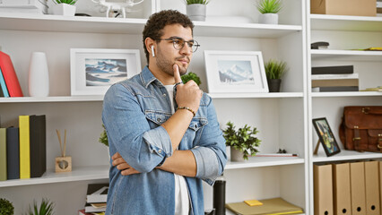 A thoughtful man with earbuds and glasses stands in an office, surrounded by shelves and plants.