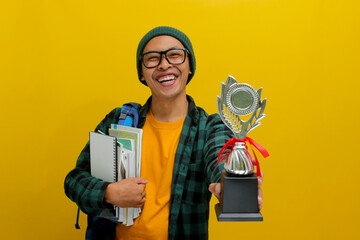 Asian student, dressed in a beanie hat and casual shirt, holds books and a trophy, celebrating the...