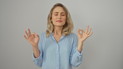 A serene blonde woman meditates in a blue shirt against a white backdrop, emanating calmness and...