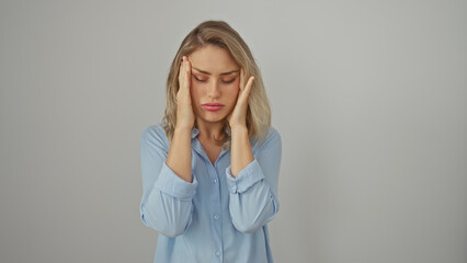 Stressed young woman with blonde hair, dressed in blue, shows headache against isolated white...
