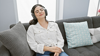 A peaceful young hispanic woman enjoys music on headphones while relaxing on a sofa in a cozy...