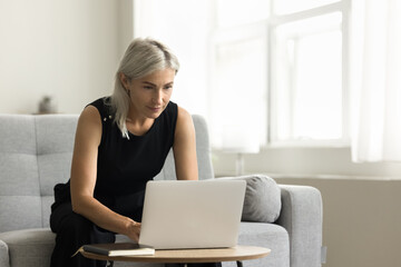 Serious senior freelance businesswoman working from home, using laptop computer at small table, sitting on couch, typing, chatting online. Retired lady shopping on Internet