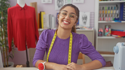 A smiling young hispanic woman with glasses and a measuring tape poses in a vibrant tailoring workshop.