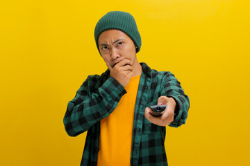 Bored young Asian man, dressed in a beanie hat and casual shirt, holds a TV remote control, flipping through different channels in search of an interesting program while watching TV, yellow background