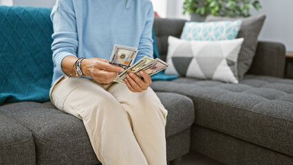 A mature woman counts us dollars sitting on a couch in a modern living room setting, implying...