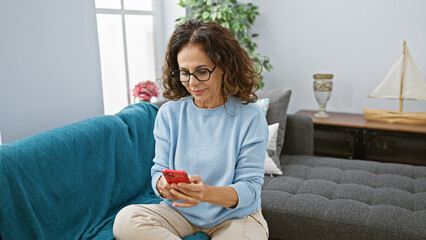 Middle-aged hispanic woman with curly hair using smartphone in cozy living room interior.