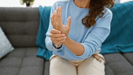 Mature hispanic woman with curly hair experiencing wrist pain at home, sitting on a grey sofa