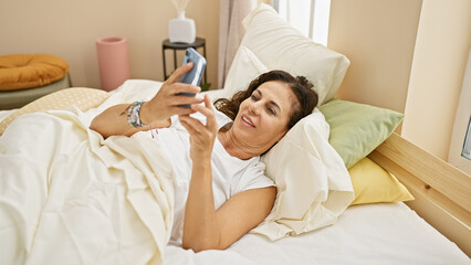 A mature hispanic woman relaxes in a bedroom, using a smartphone while lying comfortably in bed,...