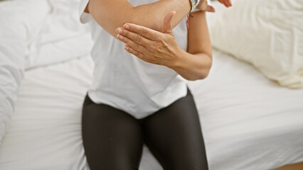 Mature hispanic woman suffering from elbow pain sitting on a bedroom's bed indoors.