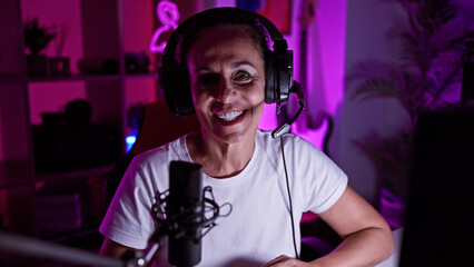 A cheerful middle-aged hispanic woman wearing headphones speaks into a microphone in a neon-lit...