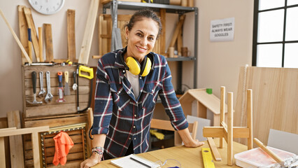 A smiling hispanic mature woman with curly hair wearing safety headphones stands at a carpentry workshop.
