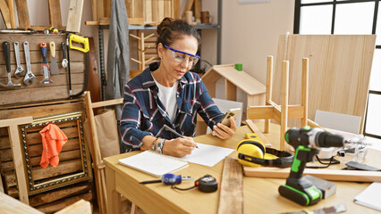Hispanic woman in glasses examines smartphone while jotting notes in a well-equipped woodworking...
