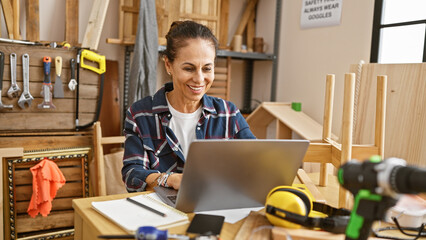 A smiling woman using a laptop in a woodworking workshop surrounded by tools and wooden pieces.