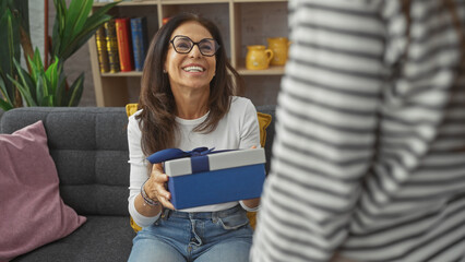 A smiling woman receives a blue gift from another in a cozy living room, depicting family affection...