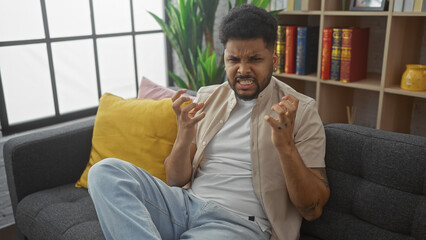 An african american man grimaces on a sofa in a modern living room, expressing frustration or...