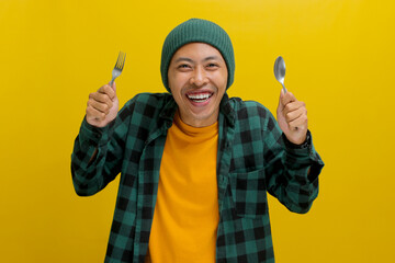Cheerful young Asian man, clad in a beanie hat and casual shirt, holds a fork and a spoon, smiling...