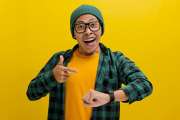 Cheerful Asian man in a beanie and casual shirt, wearing eyeglasses, gestures towards his wristwatch with a smile. Friendly reminder to keep track of time! Isolated on a yellow background