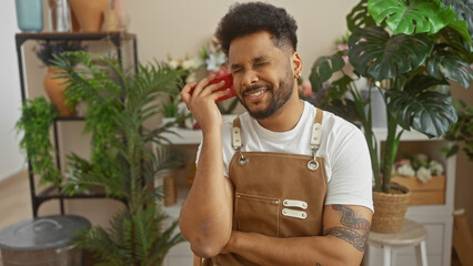 Smiling black man in apron using smartphone in flower shop, surrounded by green plants.