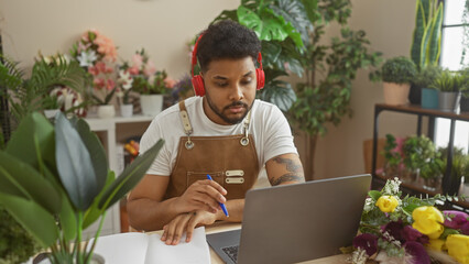 A focused man wearing headphones works on a laptop in a florist shop surrounded by plants and flowers.