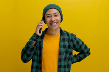 A confident Asian man, sporting a beanie hat and casual shirt, wears a smile of contentment while engaged in a phone conversation, exuding happiness as he stands against a yellow background