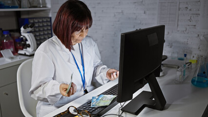 Mature woman counting chilean pesos in a laboratory setting, working on a computer indoors.