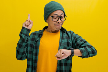 Enthusiastic Asian man in a beanie wearing eyeglasses, points upwards impatiently. He checks his...
