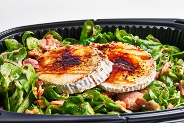 Close-up of a salad with grilled goat cheese, bacon, and spinach in a black takeout container.