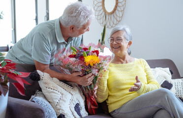 Senior man giving bouquet of flowers at his wife sitting on the sofa at home for anniversary or San...