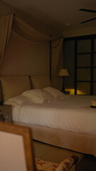Elegant hotel bedroom with unmade bed, white bedding, bedside lamp, curtain, and panama hat at night.