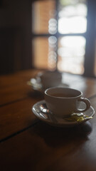 A cozy cafe scene with warm lighting featuring a white cup of coffee on a saucer with a spoon and...