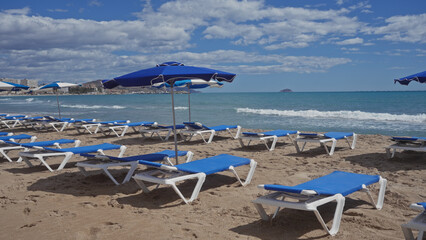 Empty blue sunbeds and umbrellas line a sandy beach with waves and clear skies hinting at a tranquil vacation setting.