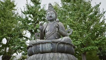 Fototapeta premium Seated buddha statue in tranquil meditation pose, enveloped by lush green trees, symbolizing peace and spirituality.