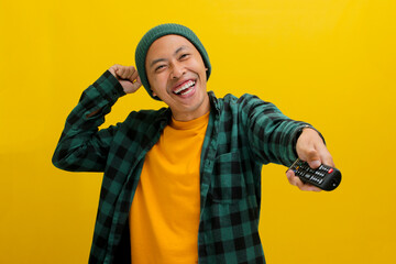 Excited Asian man, dressed in a beanie hat and casual shirt, watches a TV show or movie, pressing a button on the TV remote control while standing against a yellow background