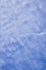 Vast blue sky with soft, textured clouds and a singular airplane trail cutting across the serene expanse.