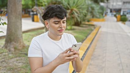 A young man uses a smartphone while sitting in a lush park with greenery and urban elements in the blurred background.