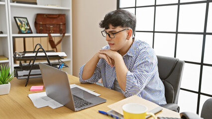 A thoughtful young man with glasses sits at a wooden desk, working on a laptop in a well-lit office.