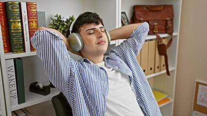 A relaxed young man wearing headphones takes a break at his modern office, surrounded by books and...