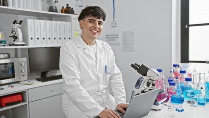 A smiling young man in a lab coat working on a laptop in a modern laboratory with a microscope and equipment.