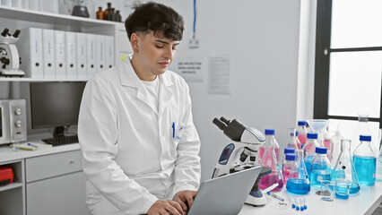 A young man in a lab coat analyzing data on a laptop in a modern laboratory with scientific equipment