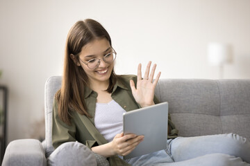 Happy pretty teenager girl in stylish eyeglasses saying hello on online video conference call, using tablet for remote Internet communication, waving greeting hand hello, smiling, laughing