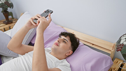 A young man relaxes in his bedroom, using his smartphone while lying on a pink pillow, evoking comfort and leisure.