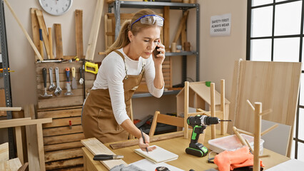 A woman carpenter multitasks talking on a phone and taking notes in a woodwork workshop.