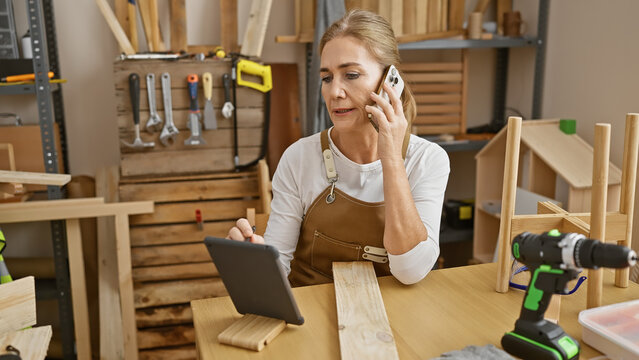 Mature blonde woman on phone while working in a carpentry workshop, surrounded by wood and tools.