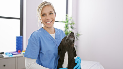 Blonde woman veterinarian smiling indoors with a loving black labrador in a clinic room.