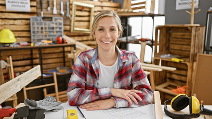 Smiling woman carpenter in plaid shirt posing in a woodshop with tools and protective gear around.